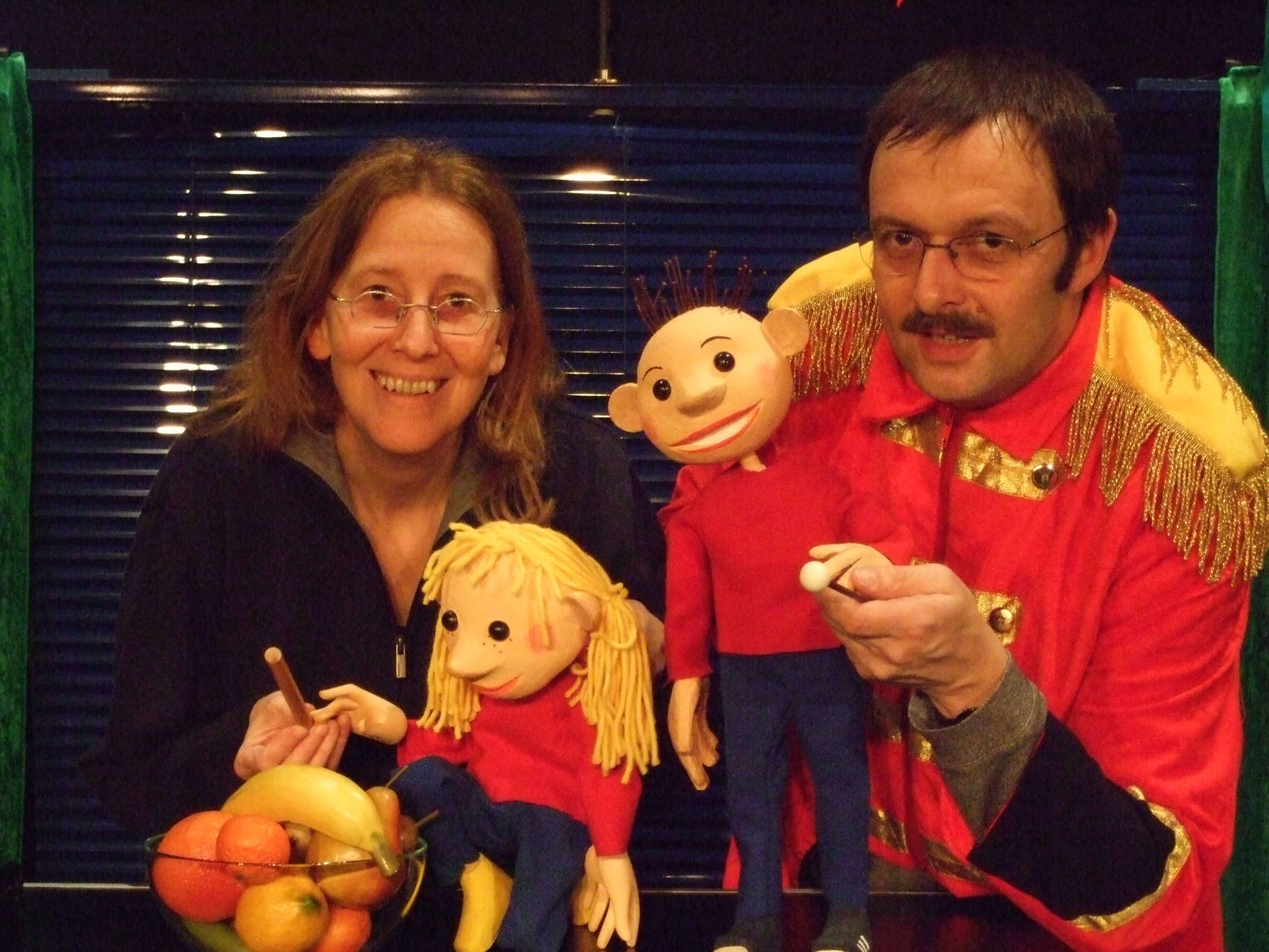 Foto: Doro Wellfonder mit der Tischfigur Flummi und Wolfgang in Zirkusdirektorjacke mit der Figur von Flo. Links daneben steht eine Schüssel mit Obst
