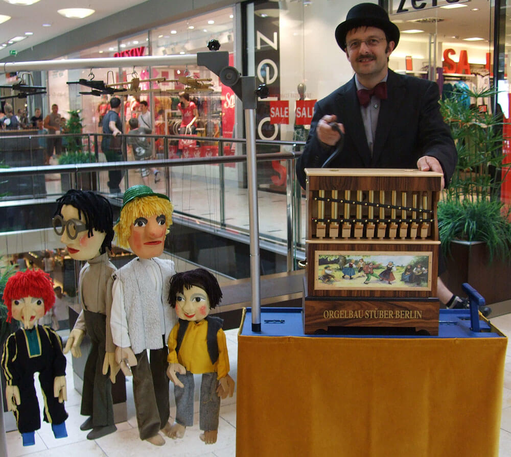 Wolfgang Kaup-Wellfonder mit der kleinen Drehorgel in einem Einkaufszentrum. Er trägt einen Hut. Daneben sieht man vier vershiedene Marionetten